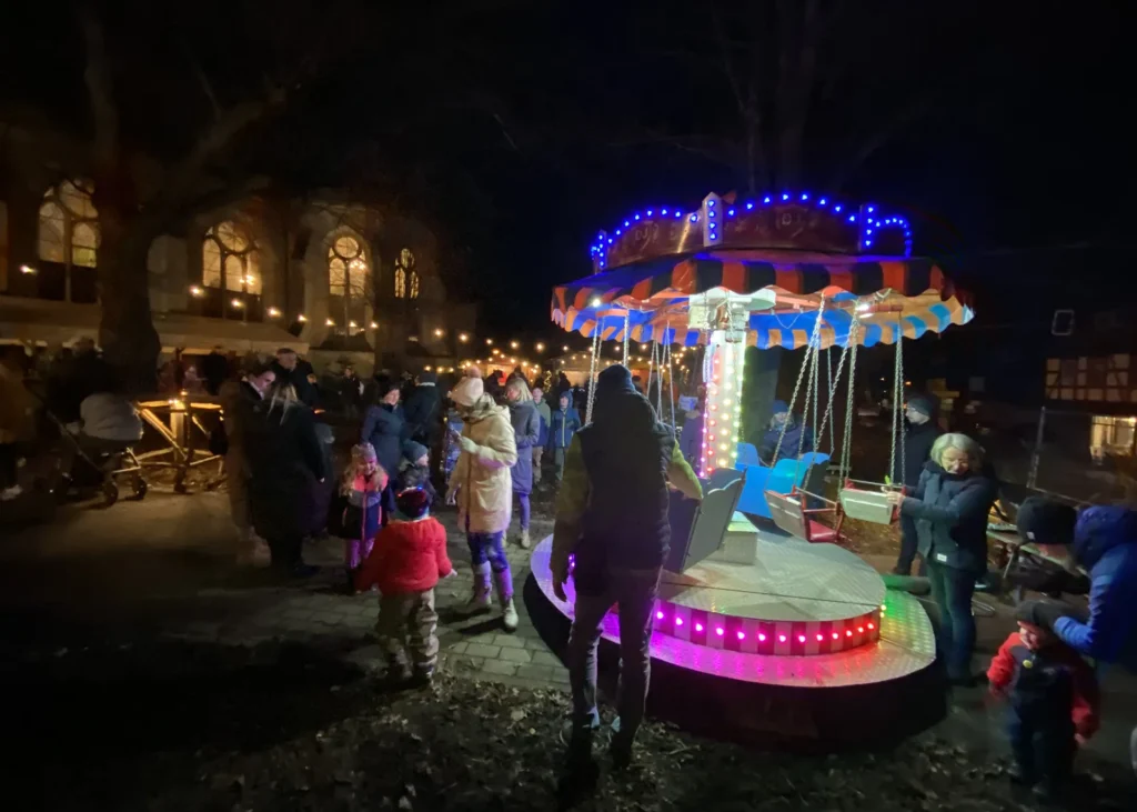 Menschen und Kinder stehen nachts an einem beleuchteten Kinderkarussell vor einem Gebäude.