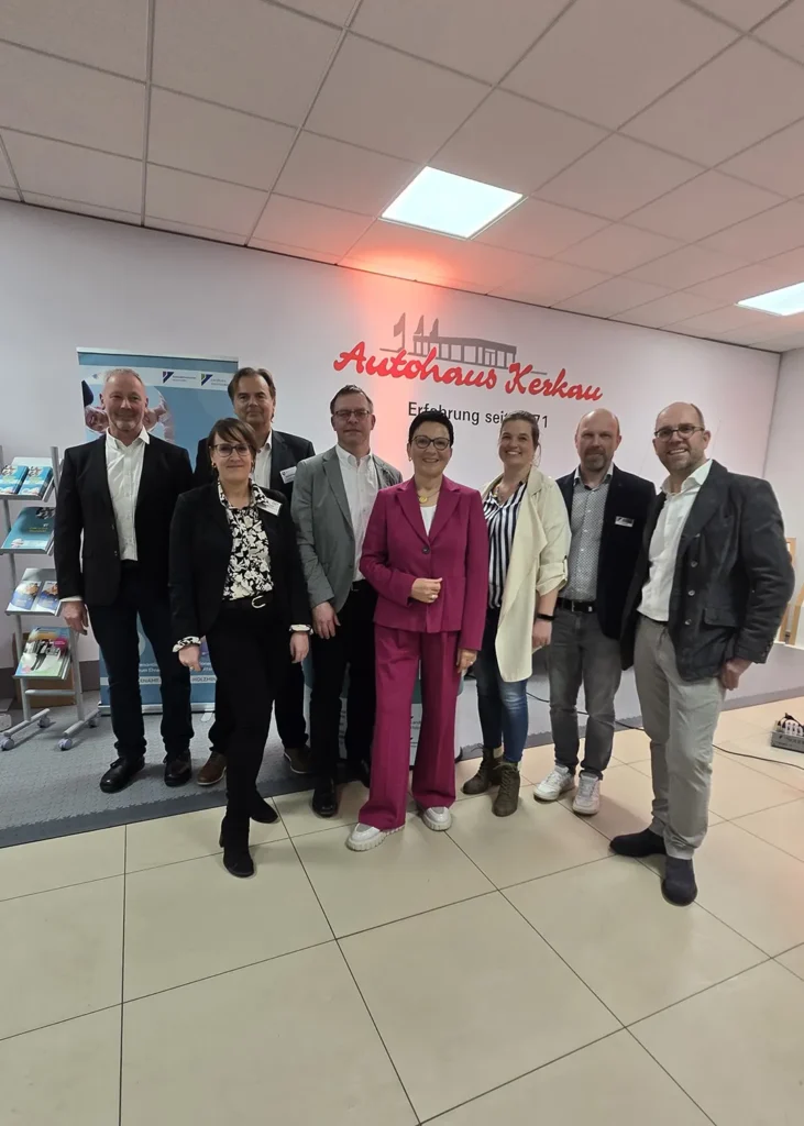 Acht Personen stehen in einem Verkaufsraum vor dem Schriftzug &bdquo;Autohaus Kerkau&ldquo;.